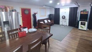 A spacious living area with a dining table, chairs, a sofa, a dartboard, and a small kitchen area with a refrigerator. - Hillbilly Haven (Equinunk)