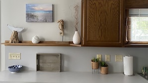 A kitchen with wooden cabinets, a shelf with decorative items, a framed picture, and a window with blinds. - The Seabreeze  (Dayton)