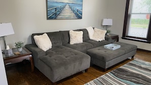 A living room with a grey sectional sofa, a matching ottoman, a striped rug, a painting of a pier, and a window with blinds. - The Seabreeze  (Dayton)