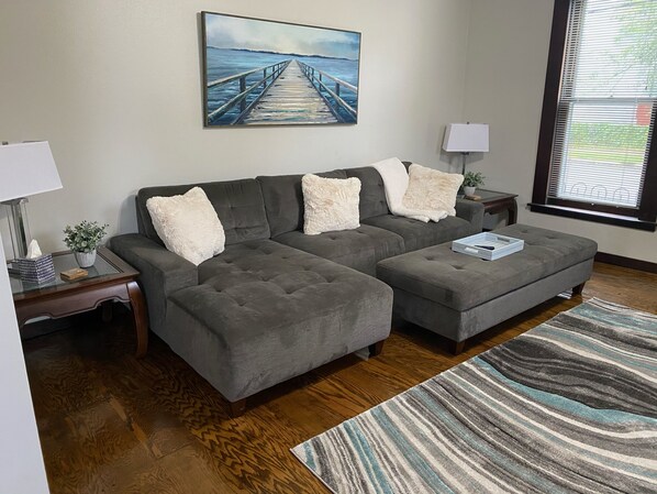A living room with a grey sectional sofa, a matching ottoman, a striped rug, a painting of a pier, and a window with blinds. - The Seabreeze (Dayton)