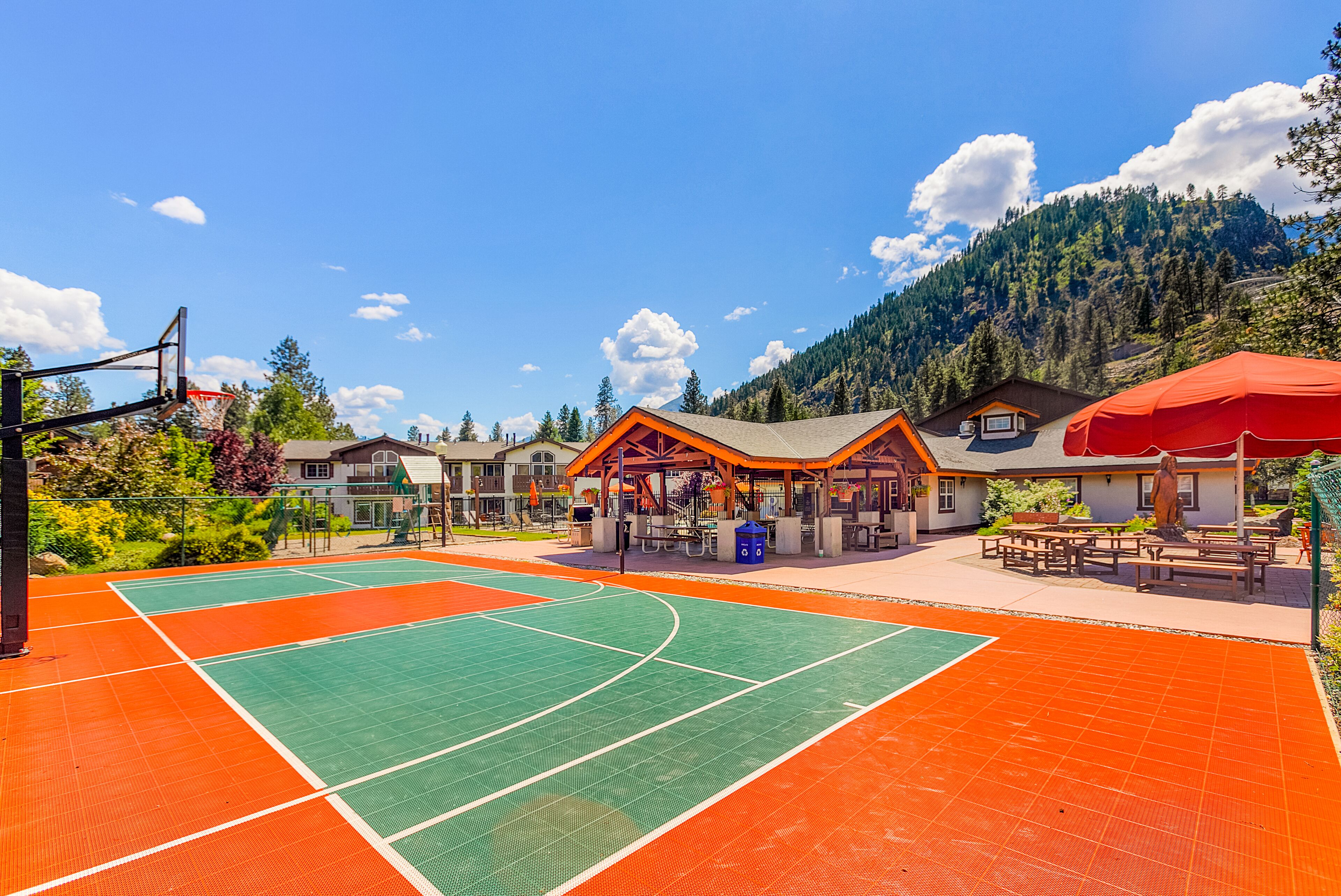 Een tennisbaan buiten met een basketbalring, picknicktafels en een overdekte zitplaats.