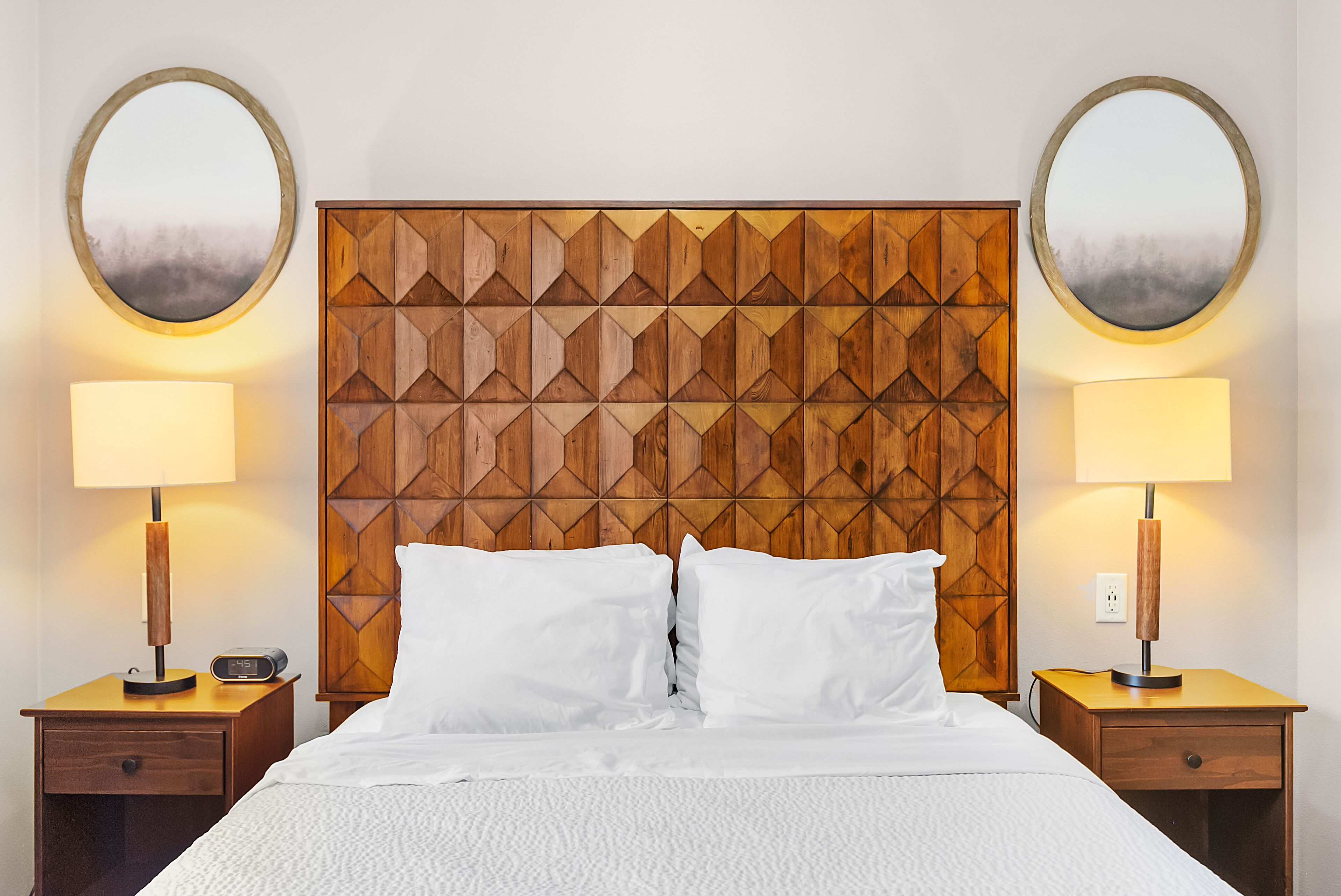 Une chambre d’hôtel avec une tête de lit en bois, deux tables de chevet, un lit avec une literie blanche et deux miroirs ronds accrochés aux murs.
