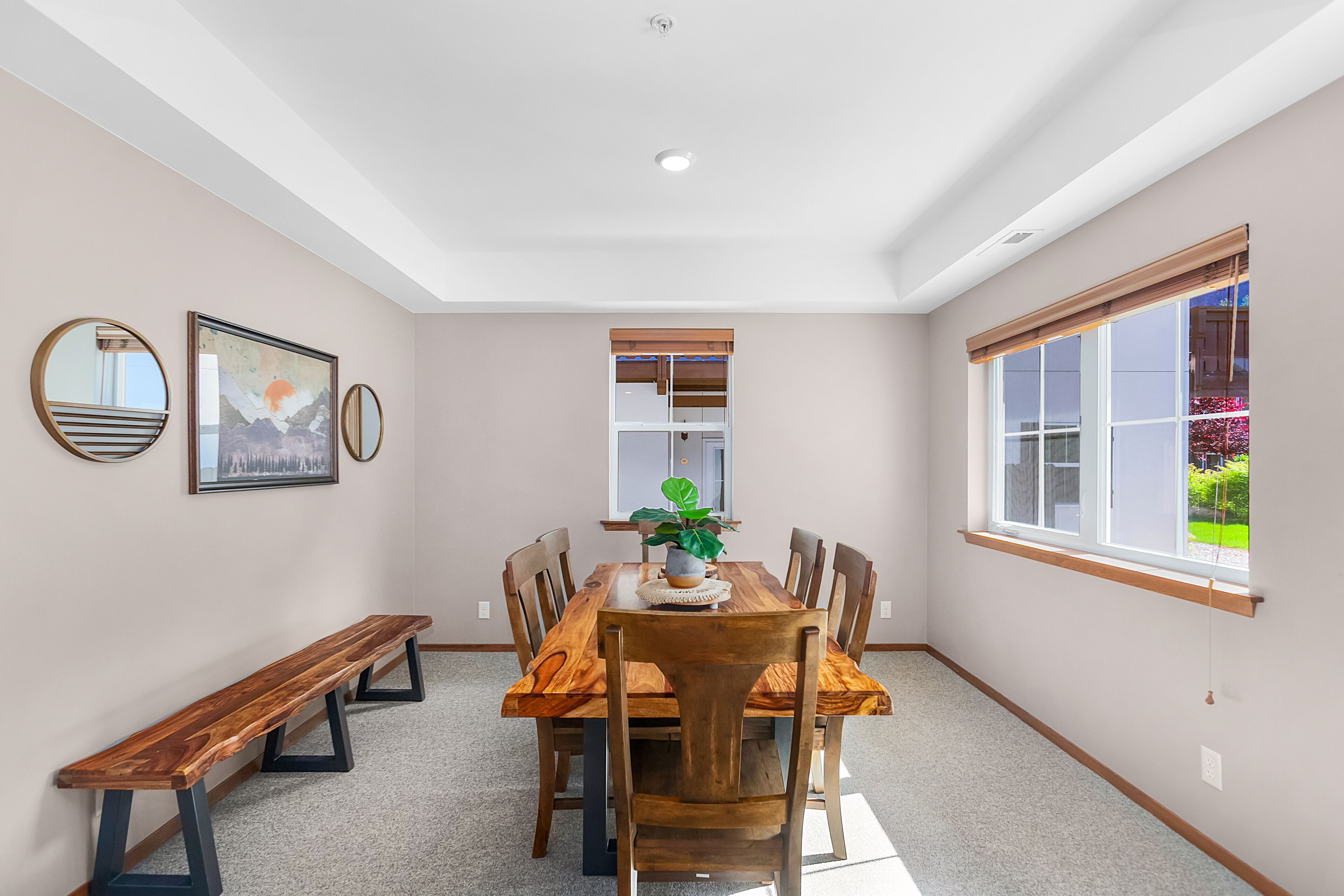 Un coin repas avec une table en bois, des chaises et un banc. Les murs sont ornés de tableaux encadrés, il y a une fenêtre avec des stores et un luminaire au plafond.