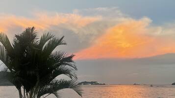 Una terraza de madera con una planta en maceta, con vista a un cuerpo de agua tranquilo y barcos a lo lejos bajo un cielo al atardecer.