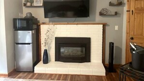 A living room with a fireplace, a flat-screen TV mounted above it, a microwave, and a refrigerator. - 'Suitest' Spot Downtown. (Anchorage)