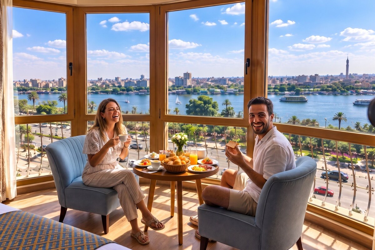 Une chambre dotée d’une grande fenêtre donnant sur un paysage urbain et sur l’eau, et comprenant deux fauteuils, une table avec un petit-déjeuner et un lit.