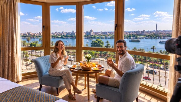A room with a large window overlooking a cityscape and water, featuring two chairs, a table with breakfast, and a bed. - Grand Bayan Hotel at Nile Plaza (Cairo)