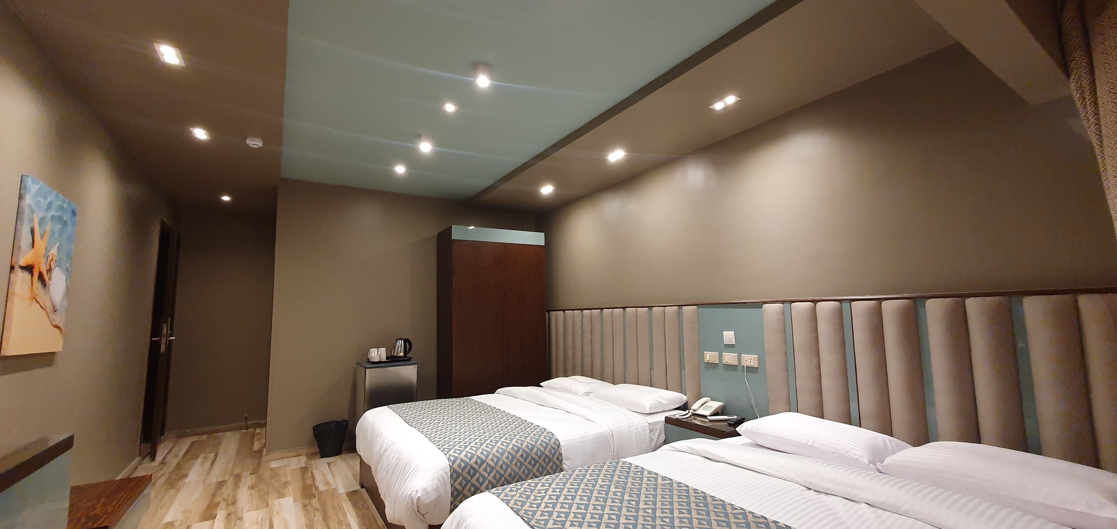Habitación de hotel con dos camas, una mesita de noche con un teléfono y una tetera, y una lámpara de pared.