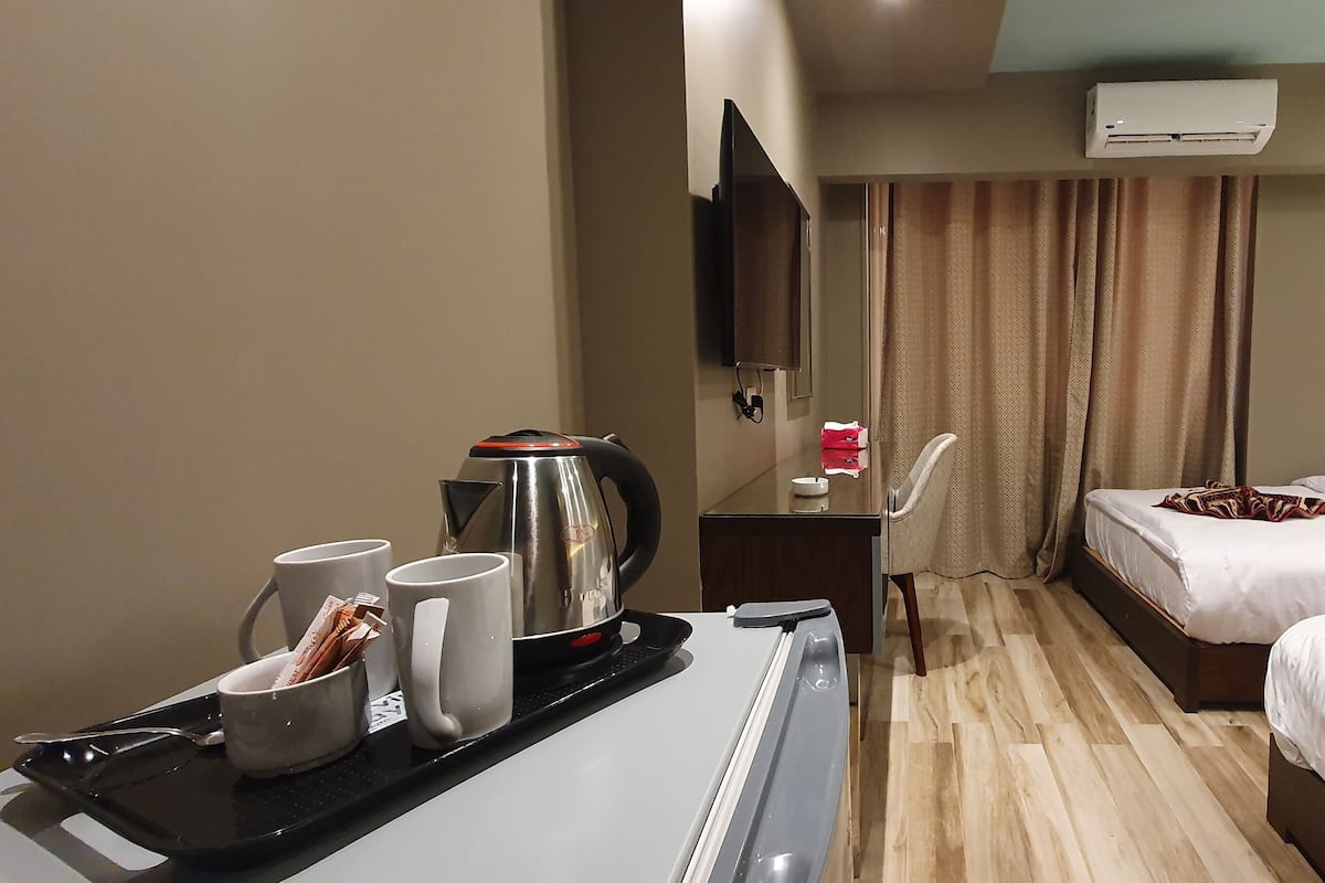 Ein Hotelzimmer mit einem Bett, einem Schreibtisch mit Wasserkocher, Tassen und Tablett, einem an der Wand montierten Fernseher und einer Klimaanlage.
