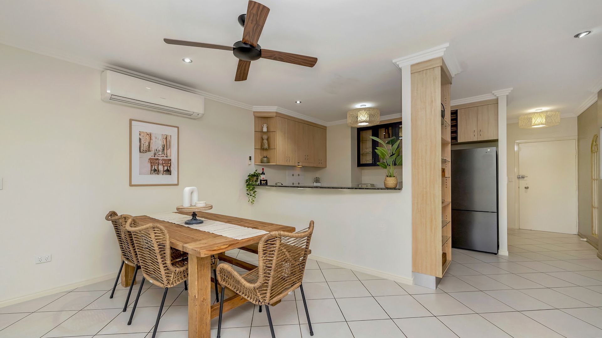 A dining area with a wooden table and chairs, a ceiling fan, and a kitchen with wooden cabinets and a refrigerator.