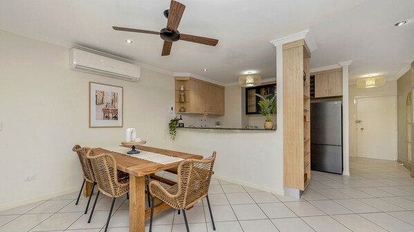 A dining area with a wooden table and chairs, a ceiling fan, and a kitchen with wooden cabinets and a refrigerator. - Apartment By The Harbour (Townsville)