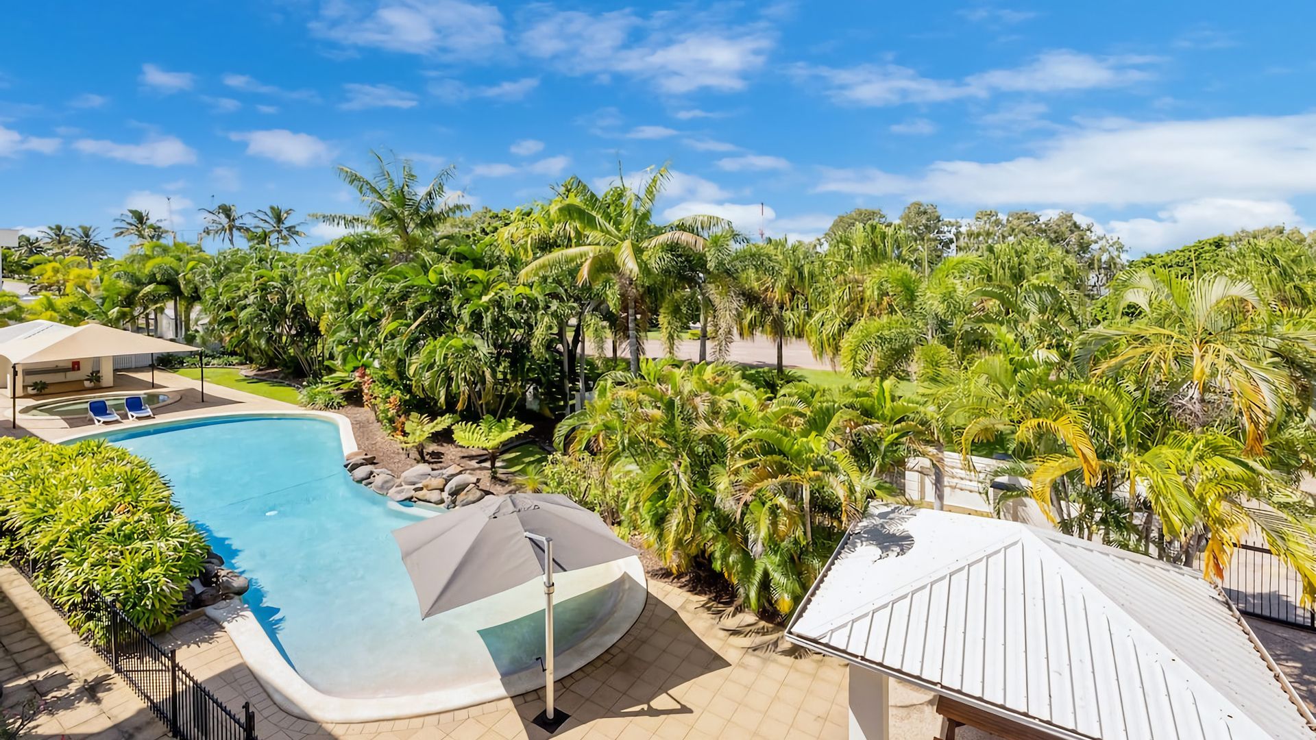 A swimming pool surrounded by lush greenery and palm trees, with a covered seating area and a white-roofed structure.