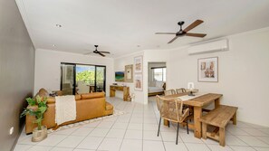 A spacious living area with a sofa, dining table, chairs, ceiling fans, and a view through a glass door. - Apartment By The Harbour (Townsville)