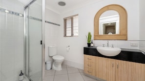 Un baño moderno con cabina de ducha de vidrio, inodoro, lavabo con espejo y ventana con persianas.