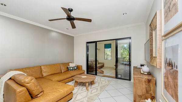 A living room with a leather sofa, a wooden coffee table, a ceiling fan, and a sliding glass door leading to an outdoor area. - Apartment By The Harbour (Townsville)