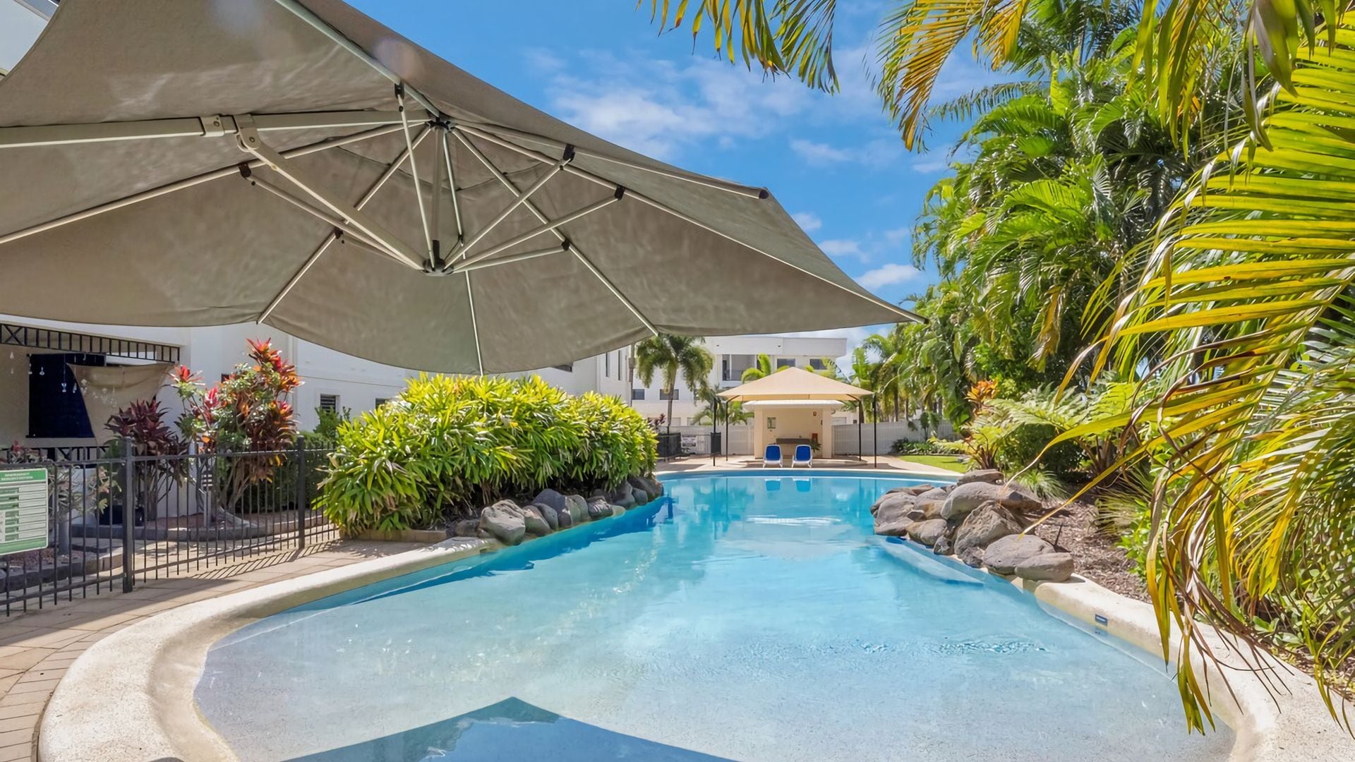 A swimming pool with a shaded area, surrounded by tropical plants and a building in the background.