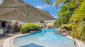 Una piscina con zona sombreada, rodeada de plantas tropicales y un edificio al fondo.