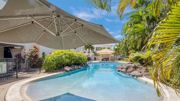 Una piscina con zona sombreada, rodeada de plantas tropicales y un edificio al fondo.