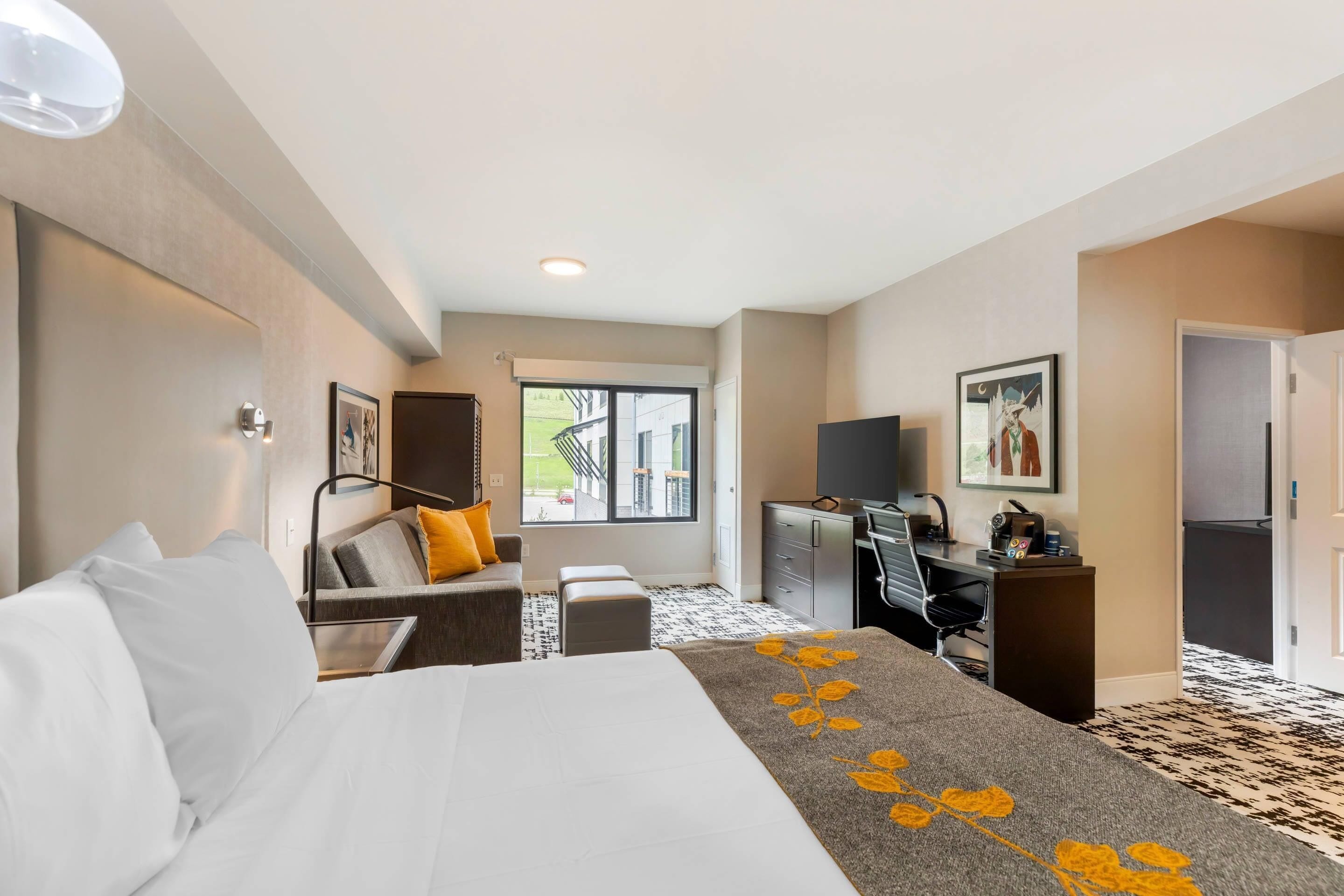 A hotel room with a bed, a desk, a chair, a sofa, a window, and a door.