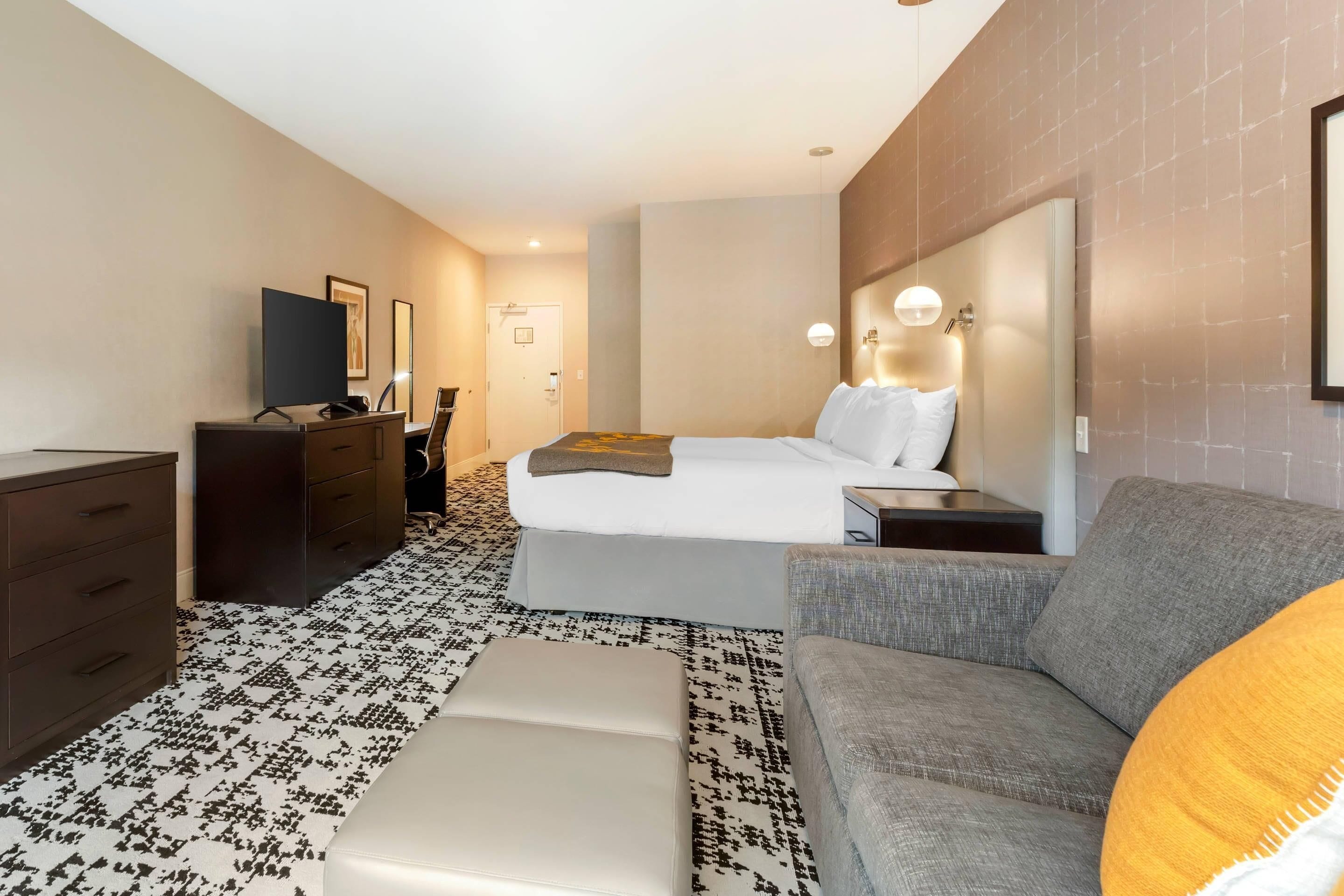 A hotel room with a bed, a gray sofa, a white ottoman, a dresser, and a TV.