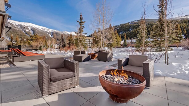 Un patio exterior con mobiliario de mimbre y una chimenea, rodeado de un paisaje nevado y montañas.