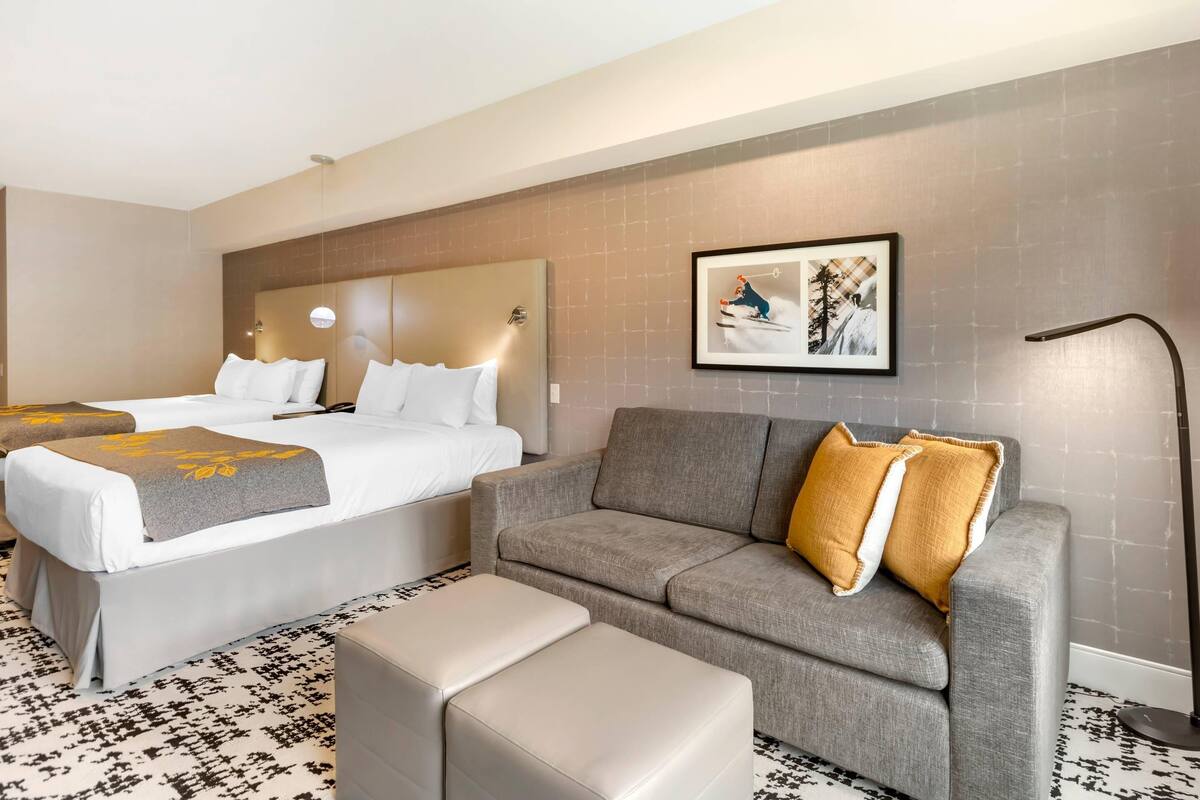 Een hotelkamer met twee bedden, een grijze bank, een kamerbreed tapijt met dessin en een ingelijst schilderij aan de muur.