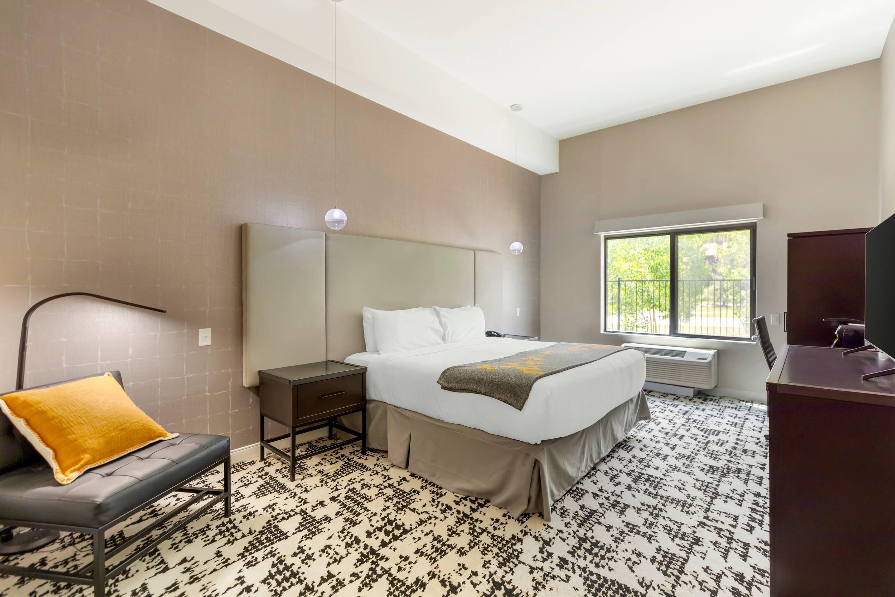 Quarto de hotel com cama, escrivaninha, cadeira, janela e radiador.