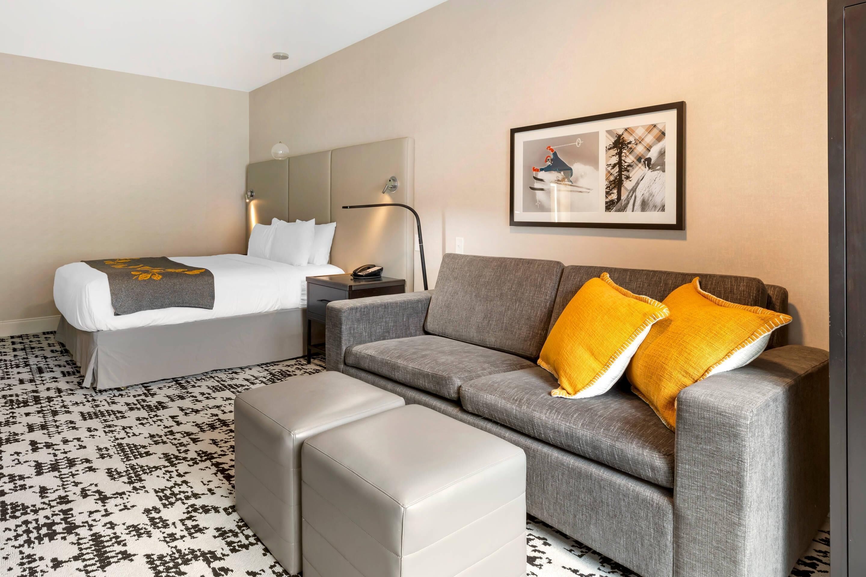 Ein Hotelzimmer mit einem Bett, einem grauen Sofa mit gelben Kissen, einem grauen Hocker, einem Nachttisch mit einer Lampe und einem gerahmten Bild an der Wand.