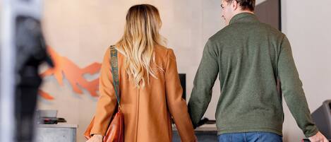 Una pareja tomándose de las manos: una mujer con abrigo marrón y un hombre con suéter verde, de pie frente a una mesa de recepción, con una maleta y un bolso negro en el suelo.