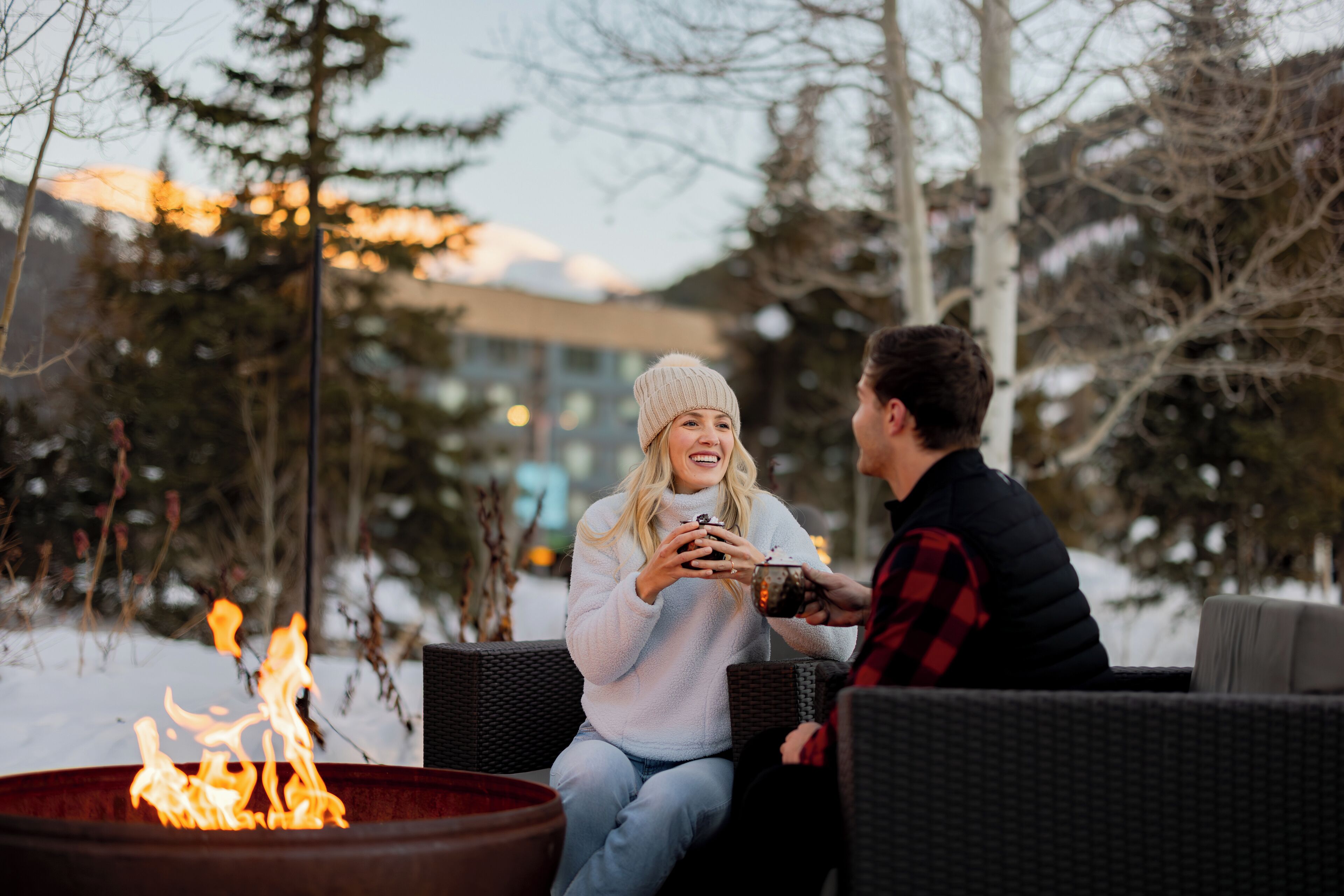 Deux personnes sont assises près d’un foyer extérieur, dans un cadre en plein air, avec de la neige au sol et des arbres en arrière-plan.