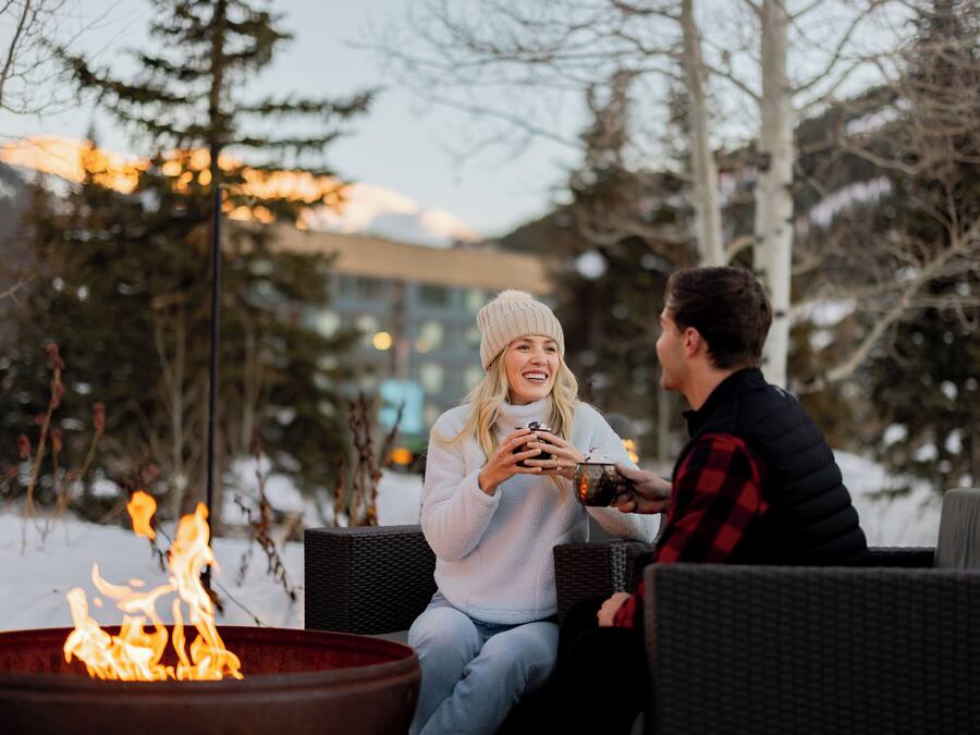 Two people sitting by a fire pit in an outdoor setting with snow on the ground and trees in the background.