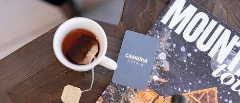 A coffee cup on a wooden table with a book titled "MOUNTAIN" and a card for "CAMBRIA hotels".