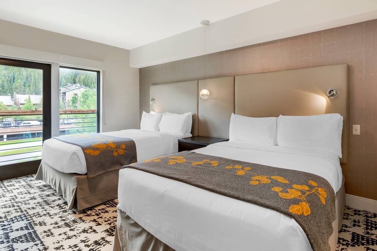 Een hotelkamer met twee bedden, een groot raam met uitzicht op een gebouw en bomen, en een kamerbreed tapijt met een patroon.