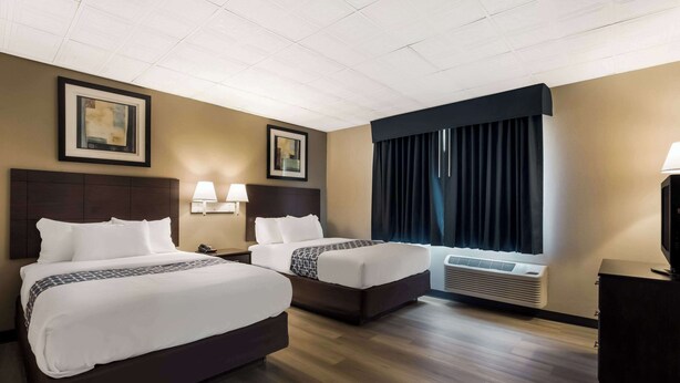 A hotel room with two beds, a large window with curtains, a wall-mounted air conditioner, and framed artwork on the walls. - Suburban Studios Millville (Millville)
