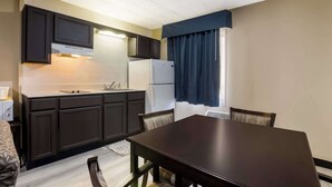 A compact kitchen with dark cabinets, a white refrigerator, and a dining area with a table and chairs. - Suburban Studios Millville (Millville)