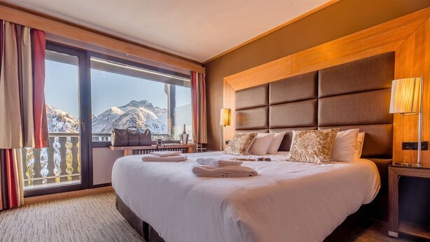 A hotel room with a large bed, a view of mountains, and a balcony with curtains.