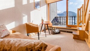 Una habitación acogedora con un ventanal grande, una silla de madera, una mesita con una botella y un bolso en el suelo.