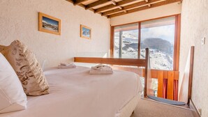 Un dormitorio con una cama, cabecera de madera, ventana con vistas a una montaña nevada y dos cuadros en la pared.