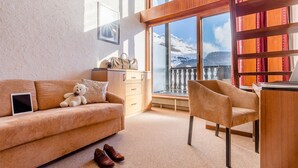 Una habitación acogedora con sofá, sillón, un osito de peluche, un par de zapatos y vistas a montañas nevadas a través de amplias ventanas.