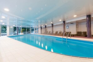 Une piscine intérieure avec des parois en verre transparent, un sol carrelé et de nombreux fauteuils de relaxation disposés le long du bord de la piscine.
