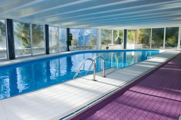 An indoor swimming pool with clear glass walls, a tiled deck, and a purple textured mat.