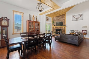 A living room with a fireplace, wooden ceiling, and a dining area with a clock and cabinet.