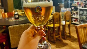 A hand holding a glass of beer with a logo, in a bar setting with tables and chairs.