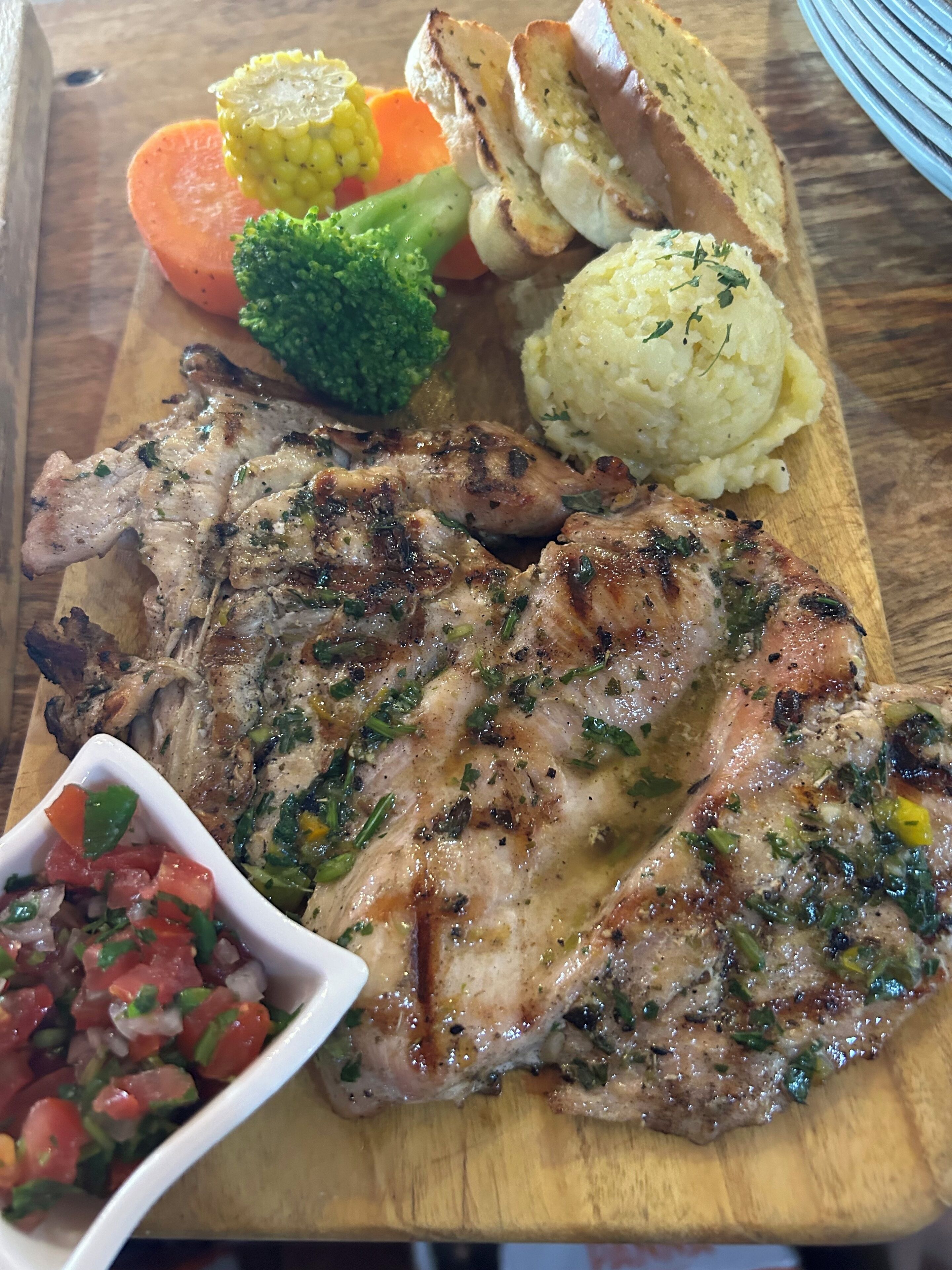 A wooden cutting board with grilled meat, vegetables, mashed potatoes, and a salsa side dish.