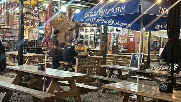 An outdoor seating area with wooden tables and benches, a blue awning with "Hofbräu München" branding, and a sign indicating a beer garden.