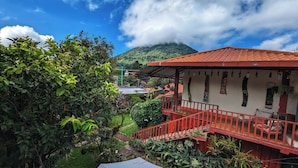 A building with a red roof and a staircase leading to a balcony. There is lush greenery surrounding the building, and a mountain is visible in the background.
