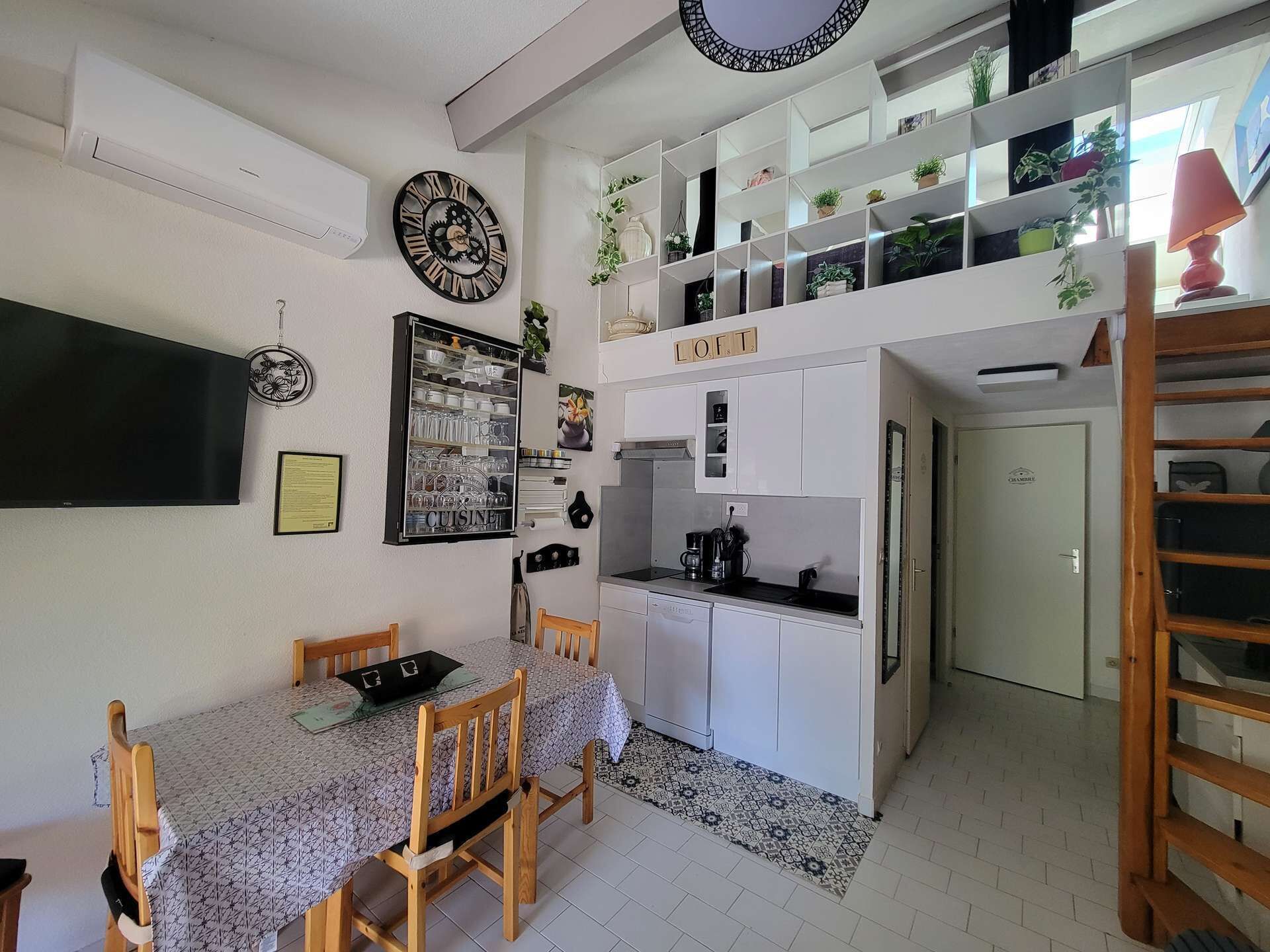 A compact kitchen with white cabinets, a dining table with chairs, a TV, and a decorative clock.