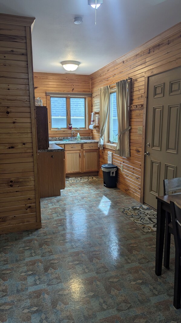 A compact kitchen with wooden walls, a sink, cabinets, a trash bin, and a door. - Cozy lake front apartment on Waldron Lake for weekly or weekend getaways. (Wawaka)