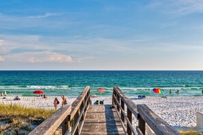 Ein hölzerner Steg führt zu einem Sandstrand mit Sonnenschirmen und Menschen.