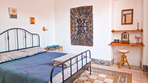 A bedroom with a bed, a small stool, a shelf with items, and a patterned rug.